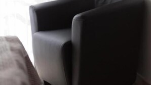 Een hoek van een kamer met een zwarte stoel, een raam met gordijnen en een bed met een deken.