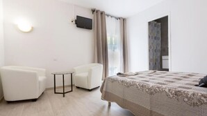 Hotelkamer met een bed, twee witte fauteuils, een klein rond tafeltje, een aan de muur bevestigde televisie en een raam met gordijnen.