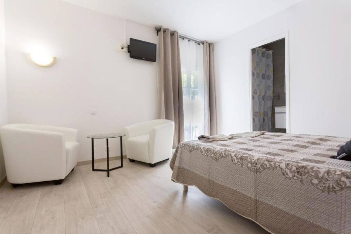 Ein Hotelzimmer mit einem Bett, zwei weißen Sesseln, einem kleinen runden Tisch, einem an der Wand montierten Fernseher und einem fenster mit Vorhängen.