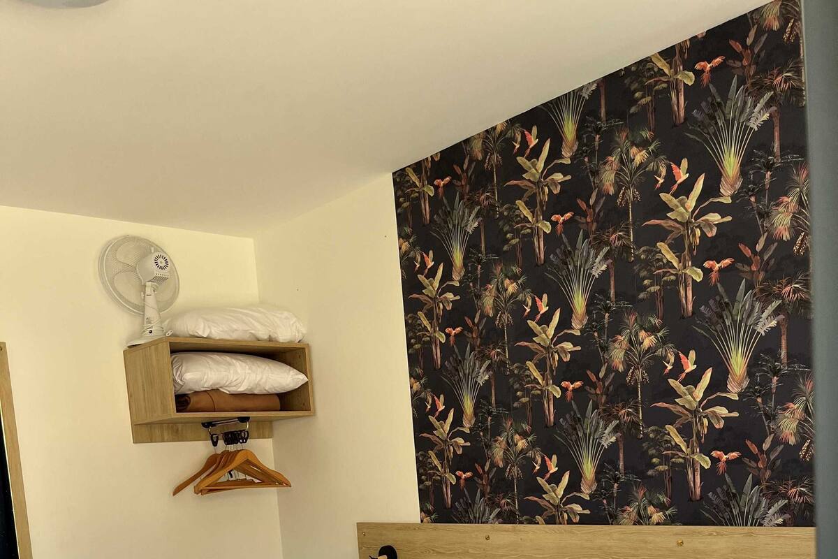 Habitació d'hotel amb un llit, un ventilador fixat a la paret, una estant amb roba doblegada i un paper pintat amb motius tropicals.