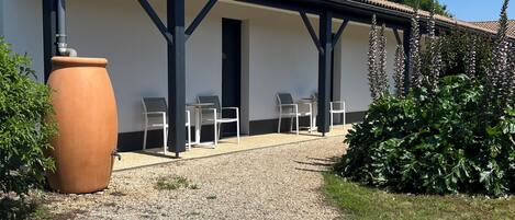 A building with a tiled roof, a gravel path leading to a covered seating area with chairs, and a large terracotta pot.