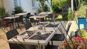 Un patio al aire libre con mesas y sillas, rodeado de plantas en macetas y un paraguas grande que proporciona sombra.