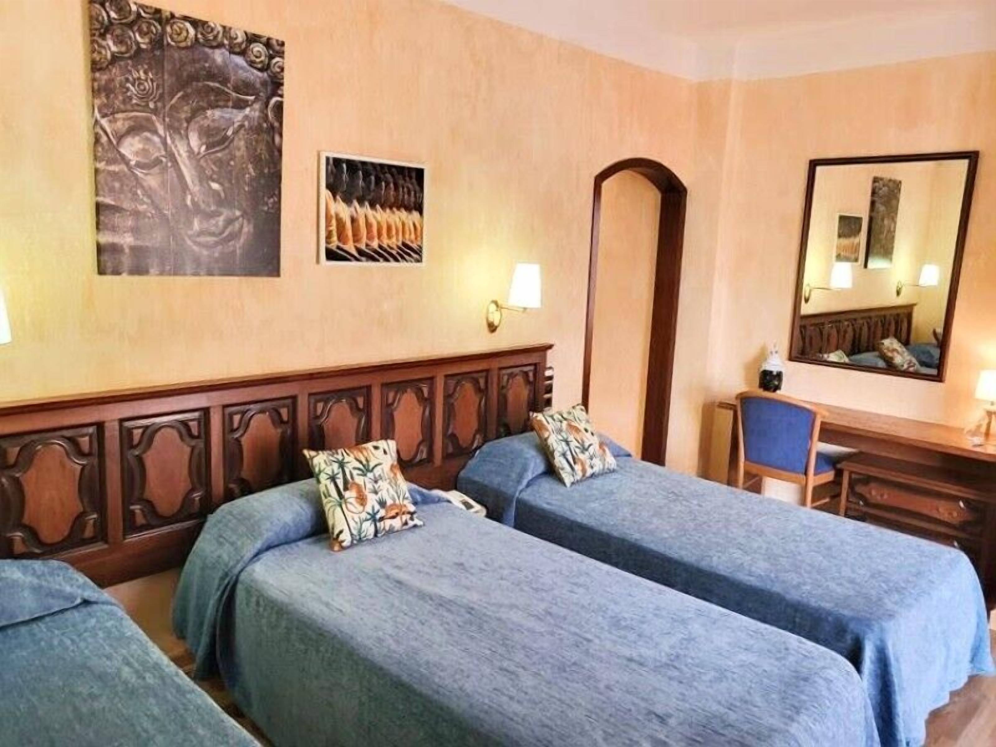 A hotel room with two single beds, a desk with a chair, a mirror, and a wall-mounted artwork.