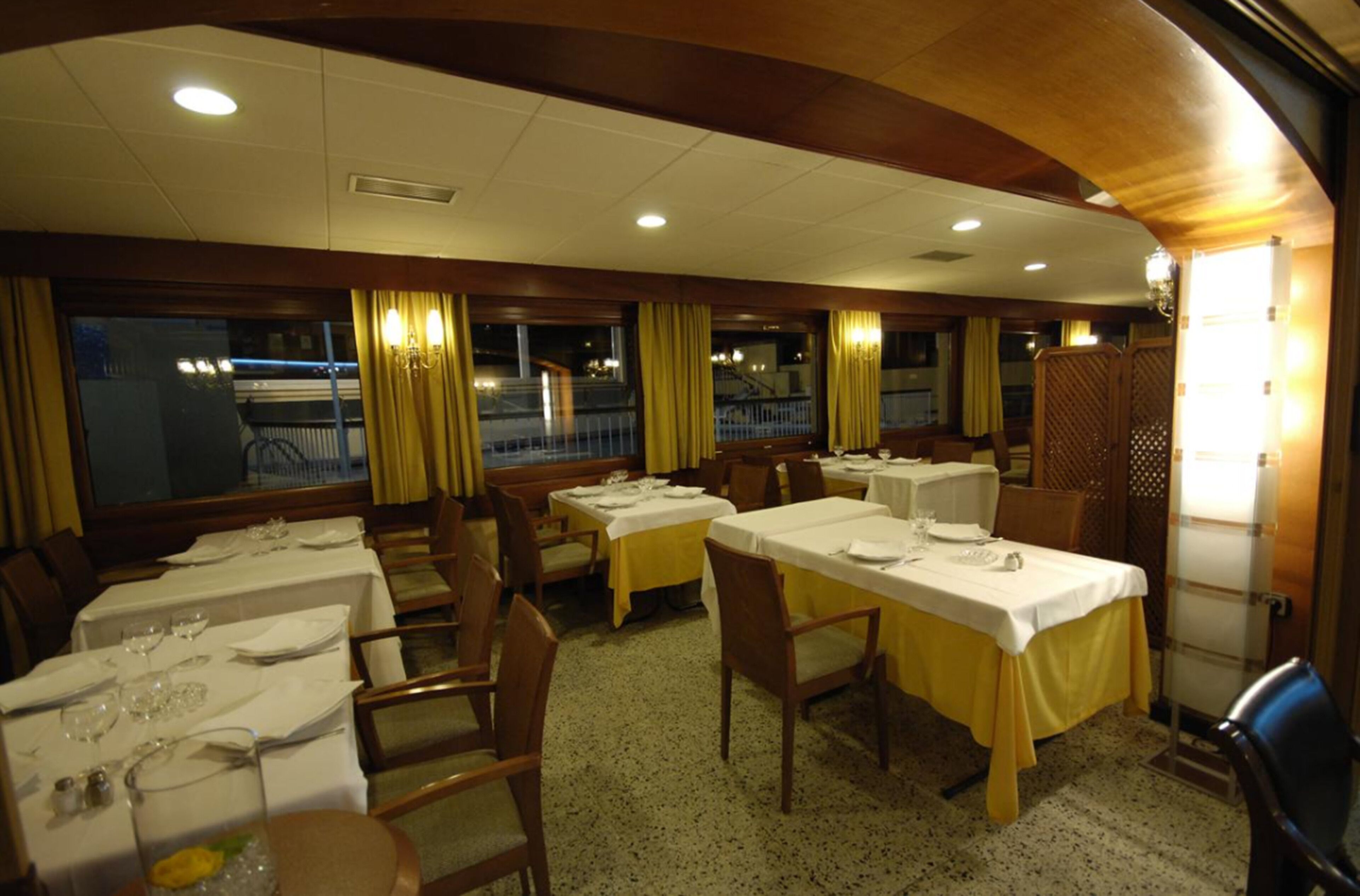 A dining area with tables set for service, wooden chairs, and a view of the outside through large windows.