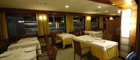 A dining area with tables set for service, wooden chairs, and a view of the outside through large windows.