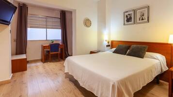 Une chambre d’hôtel avec un lit, un bureau, une chaise, une télévision, une horloge et deux tableaux encadrés au mur.