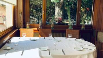 Un comedor con mesas redondas preparadas para dos comensales, sillas de madera y grandes ventanales que ofrecen vistas al exterior.