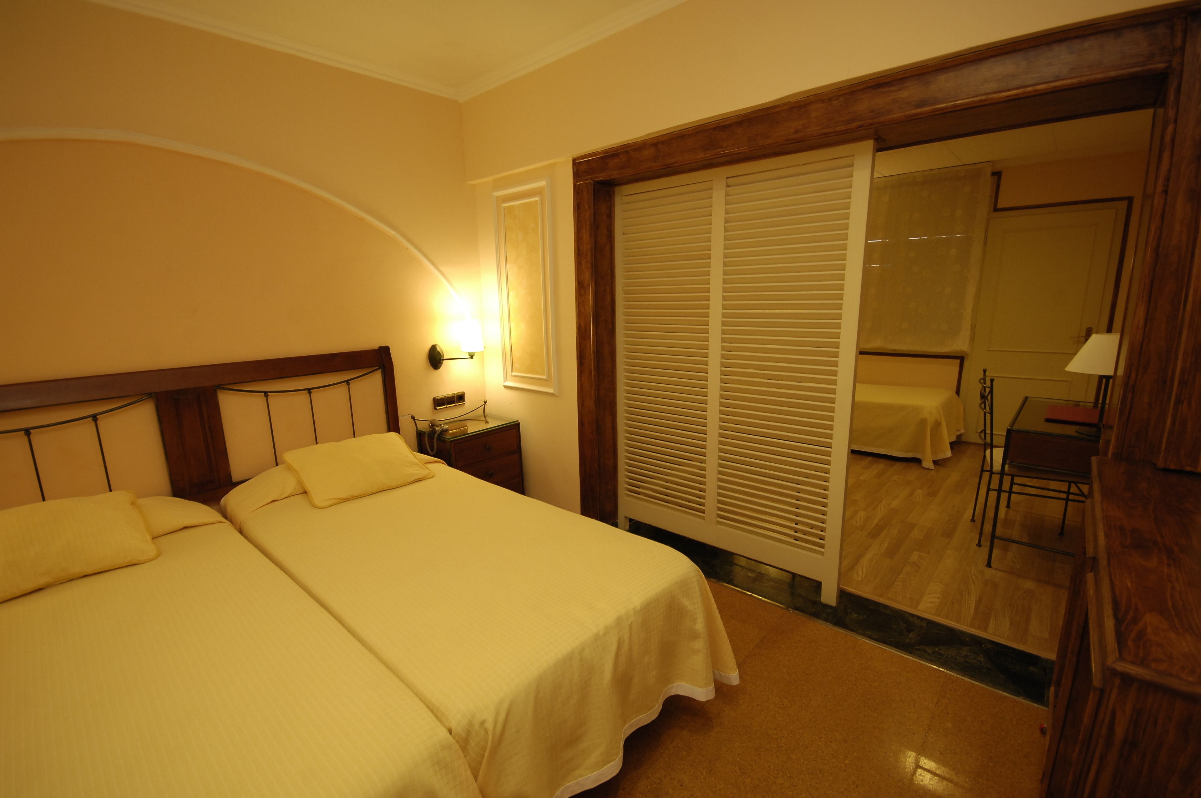 A hotel room with two beds, a nightstand, a lamp, a sliding door leading to a bathroom, and a small table with a chair.