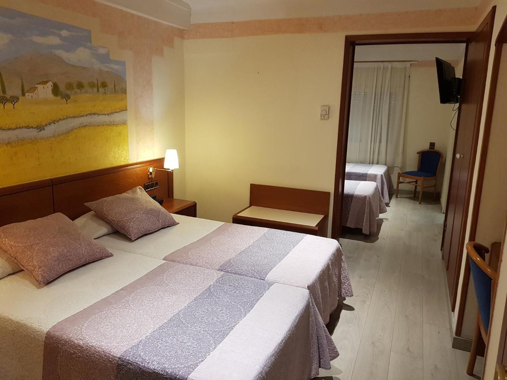Habitación de hotel con dos camas, una mesita de noche con lámpara, un cuadro en la pared y un televisor en la esquina.