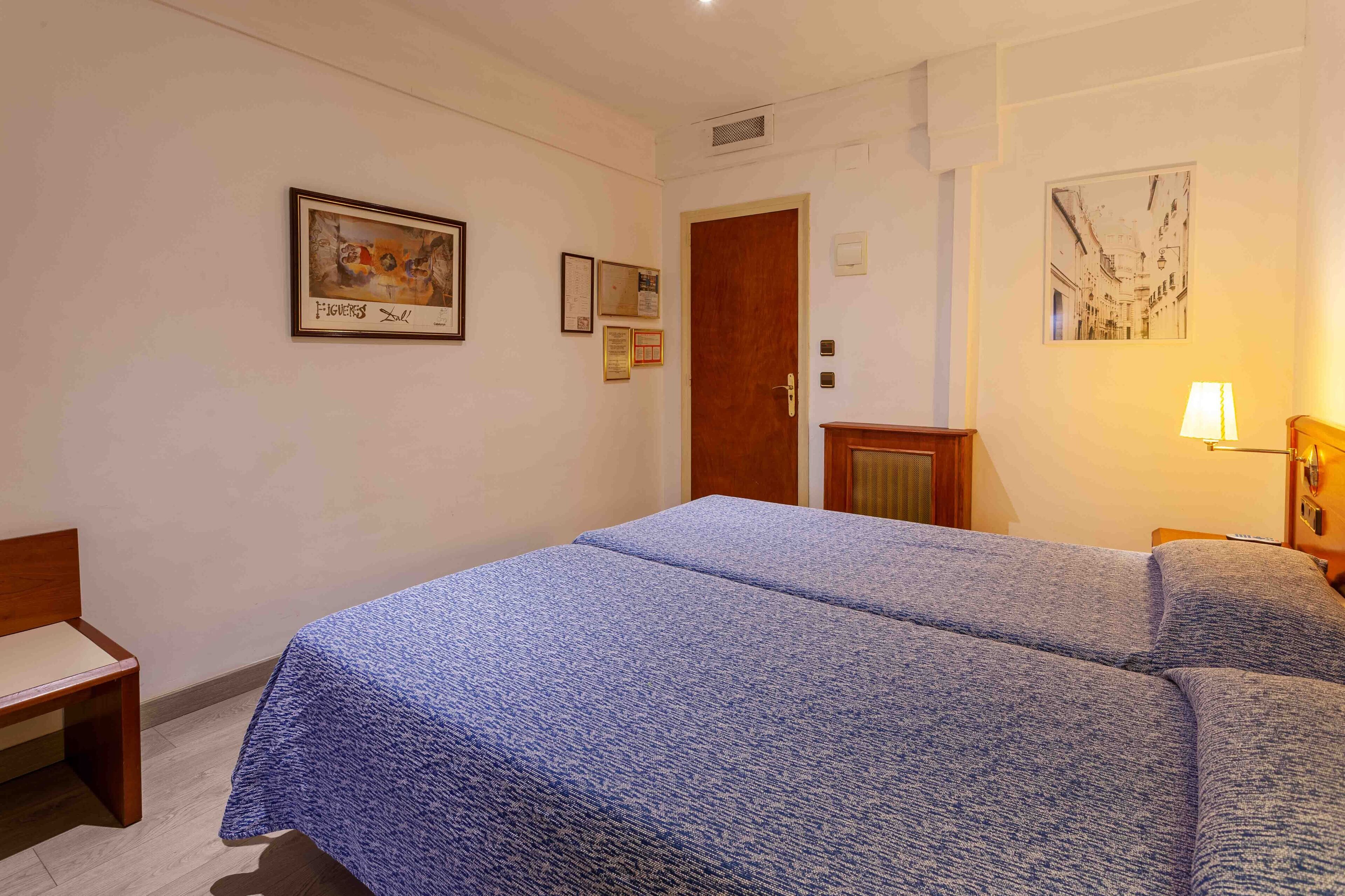 Camera d'albergo con due letti, una sedia di legno, una scrivania e una lampada.