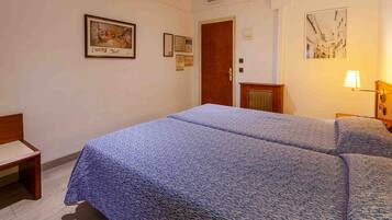 Camera d'albergo con due letti, una sedia di legno, una scrivania e una lampada.