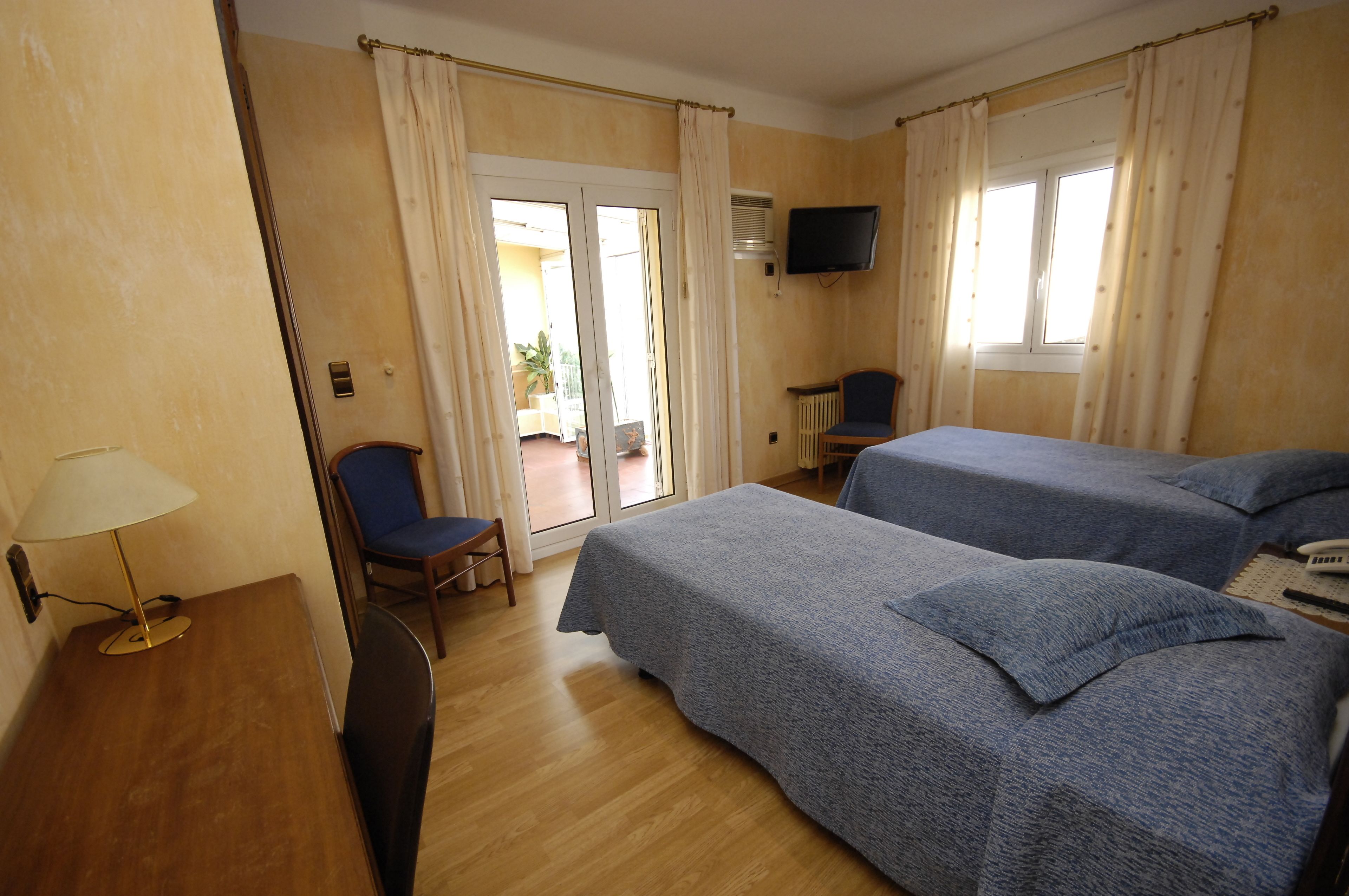 Habitación de hotel con dos camas, un escritorio con lámpara, una silla, televisión y vistas al balcón exterior.