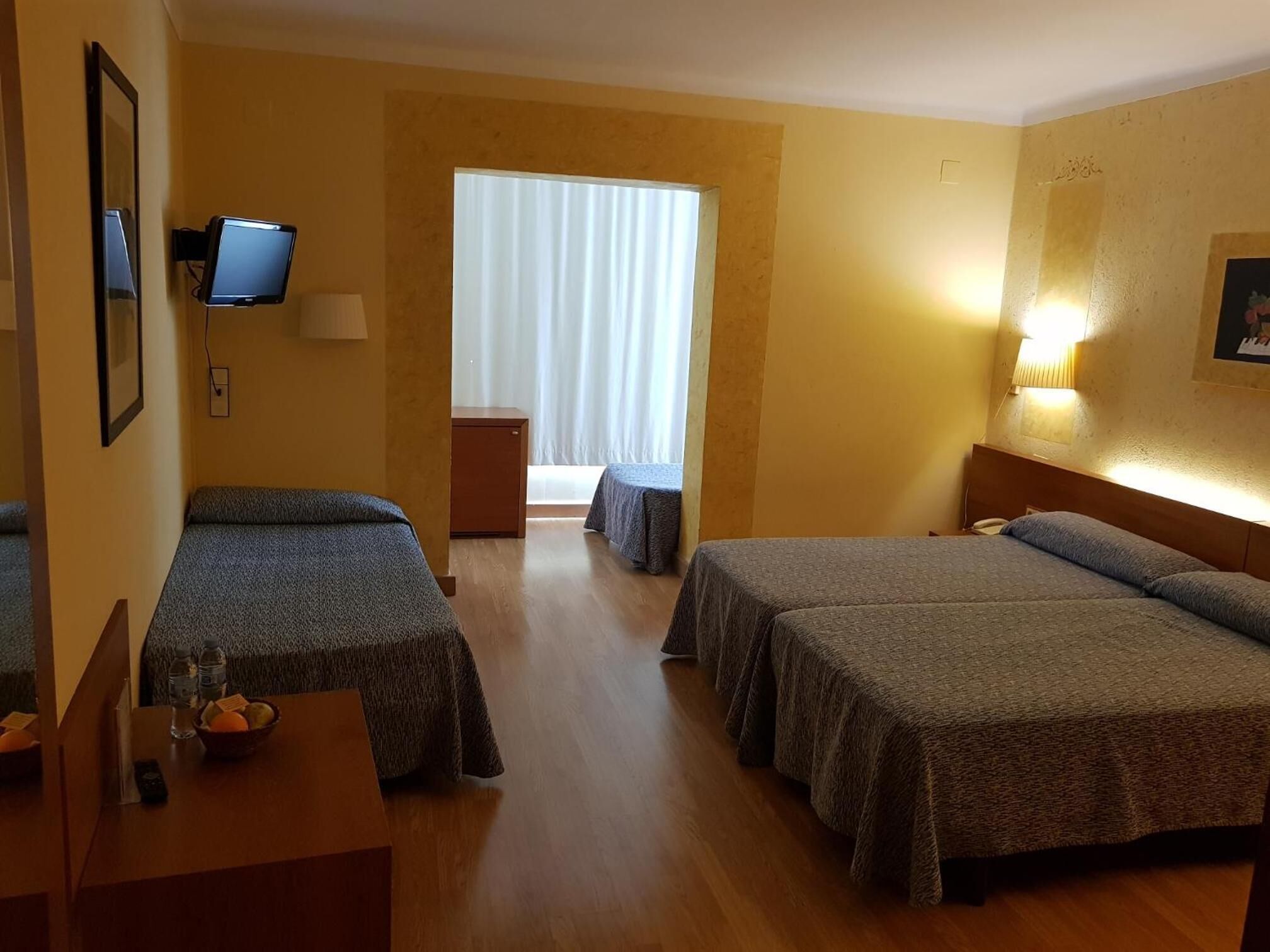 Ein Hotelzimmer mit zwei Einzelbetten, einem Schreibtisch mit Stuhl, einem kleinen Tisch mit einer Flasche und einer Fruchtschale, einem an der Wand montierten Fernseher und einem Fenster mit Vorhängen.