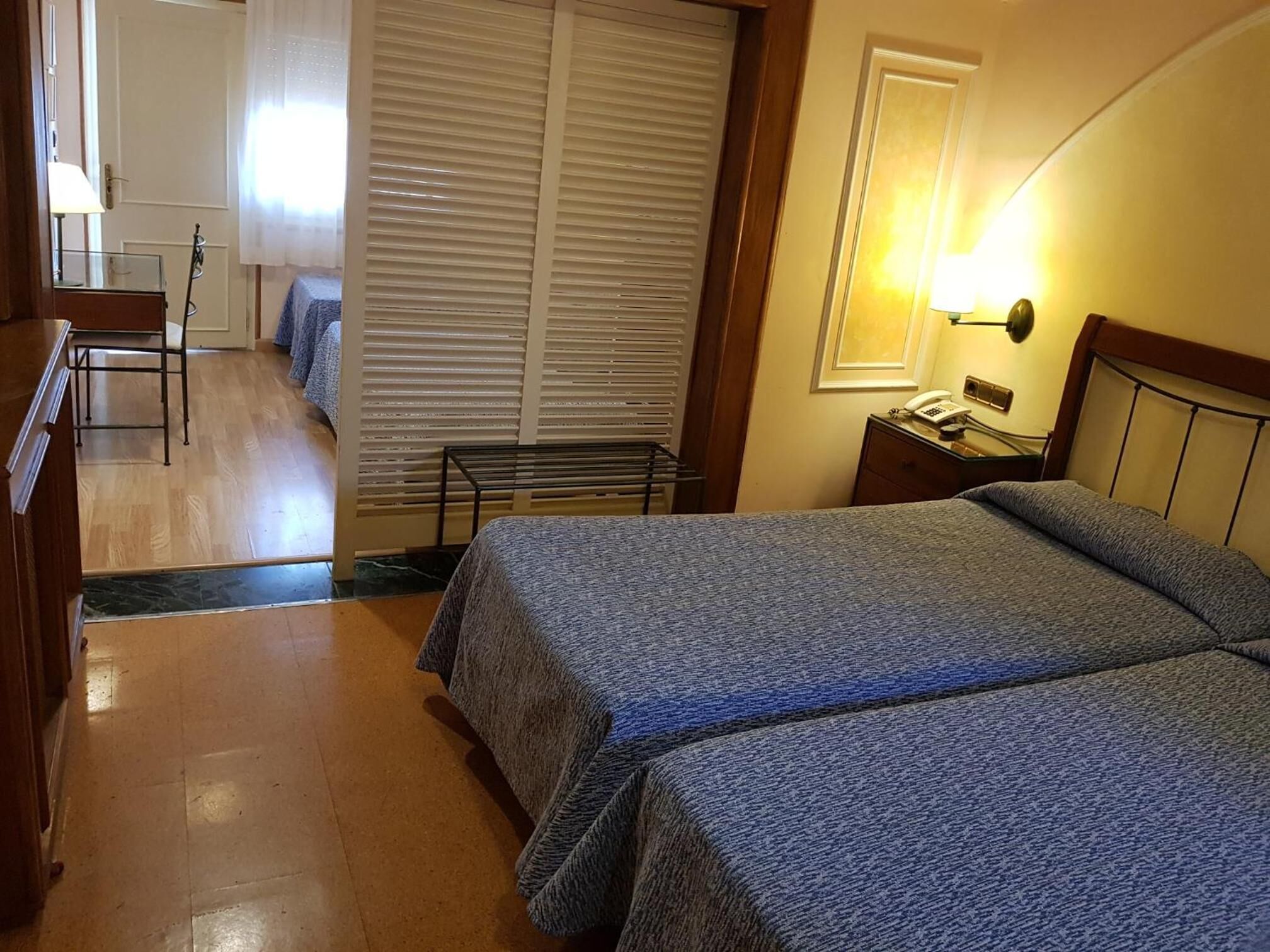Una habitación de hotel con dos camas, un escritorio, una silla, una lámpara y una ventana con persianas.