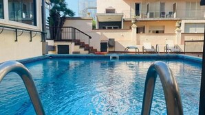 Piscina com escada de metal, rodeada por edifícios com varandas e céu claro.