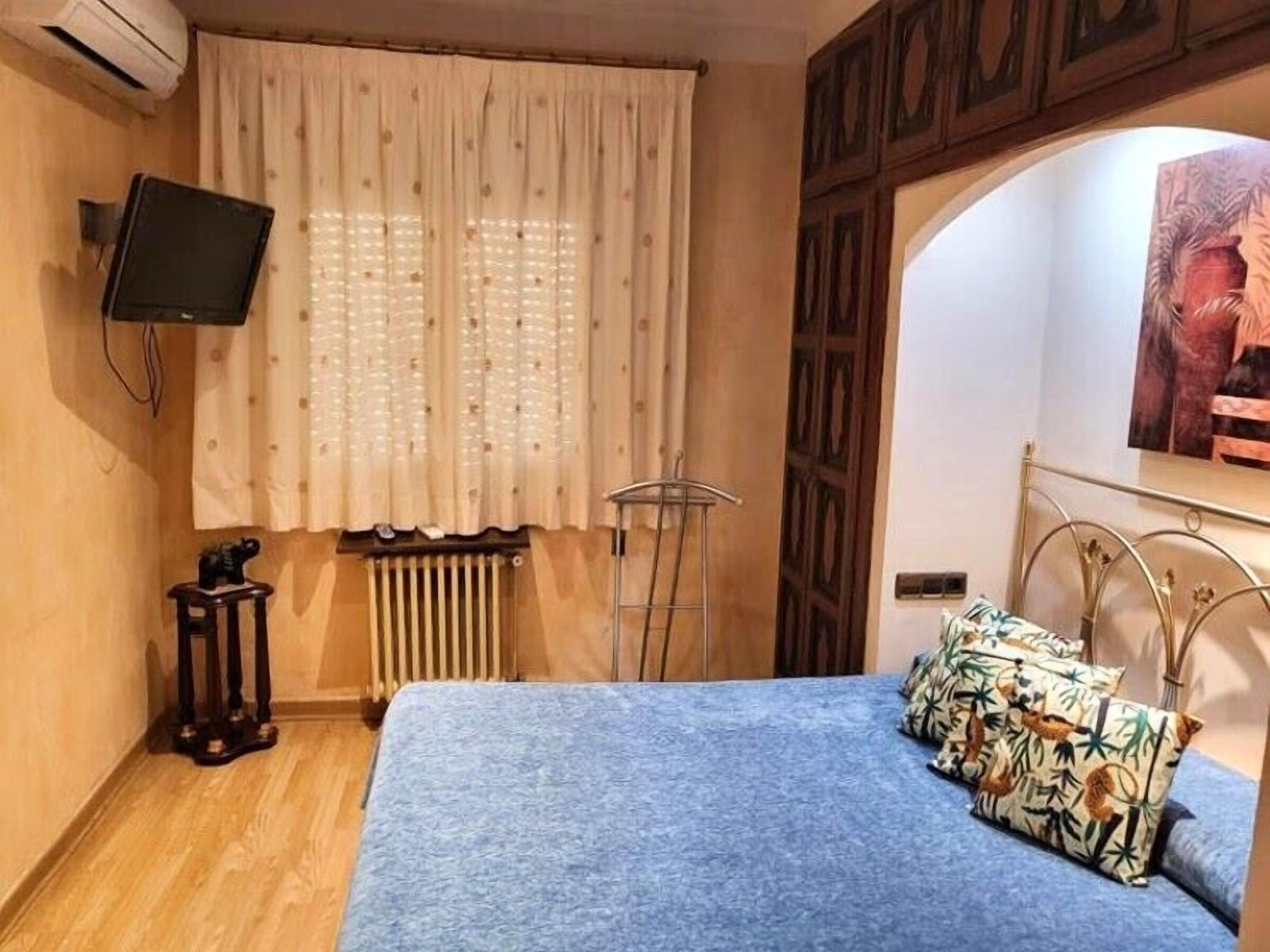 Un dormitori amb un llit, un radiador, un televisor fixat a la paret i una finestra amb cortines.