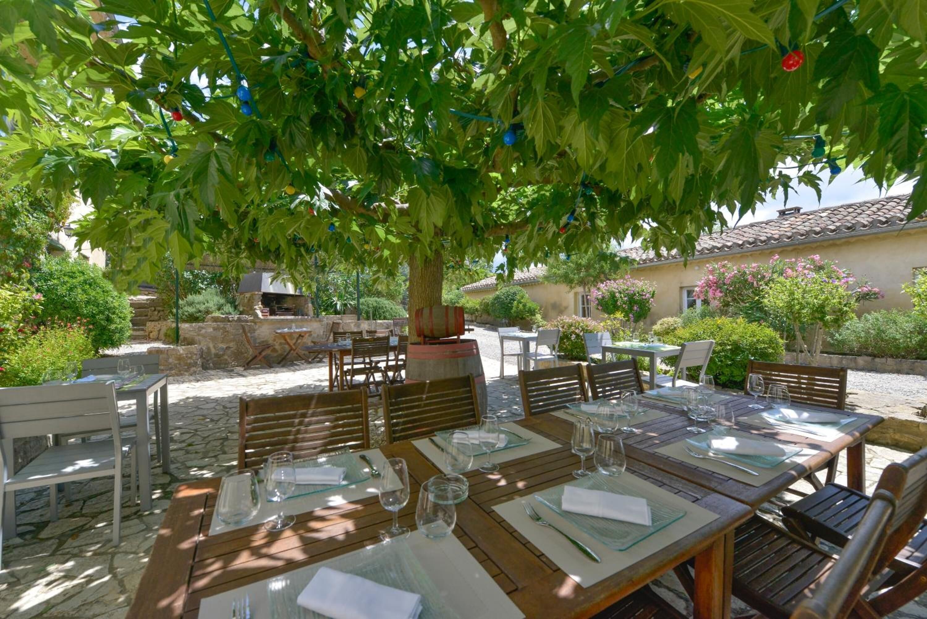 An outdoor dining area with tables and chairs under a tree, stone flooring, and a stone wall in the background.
