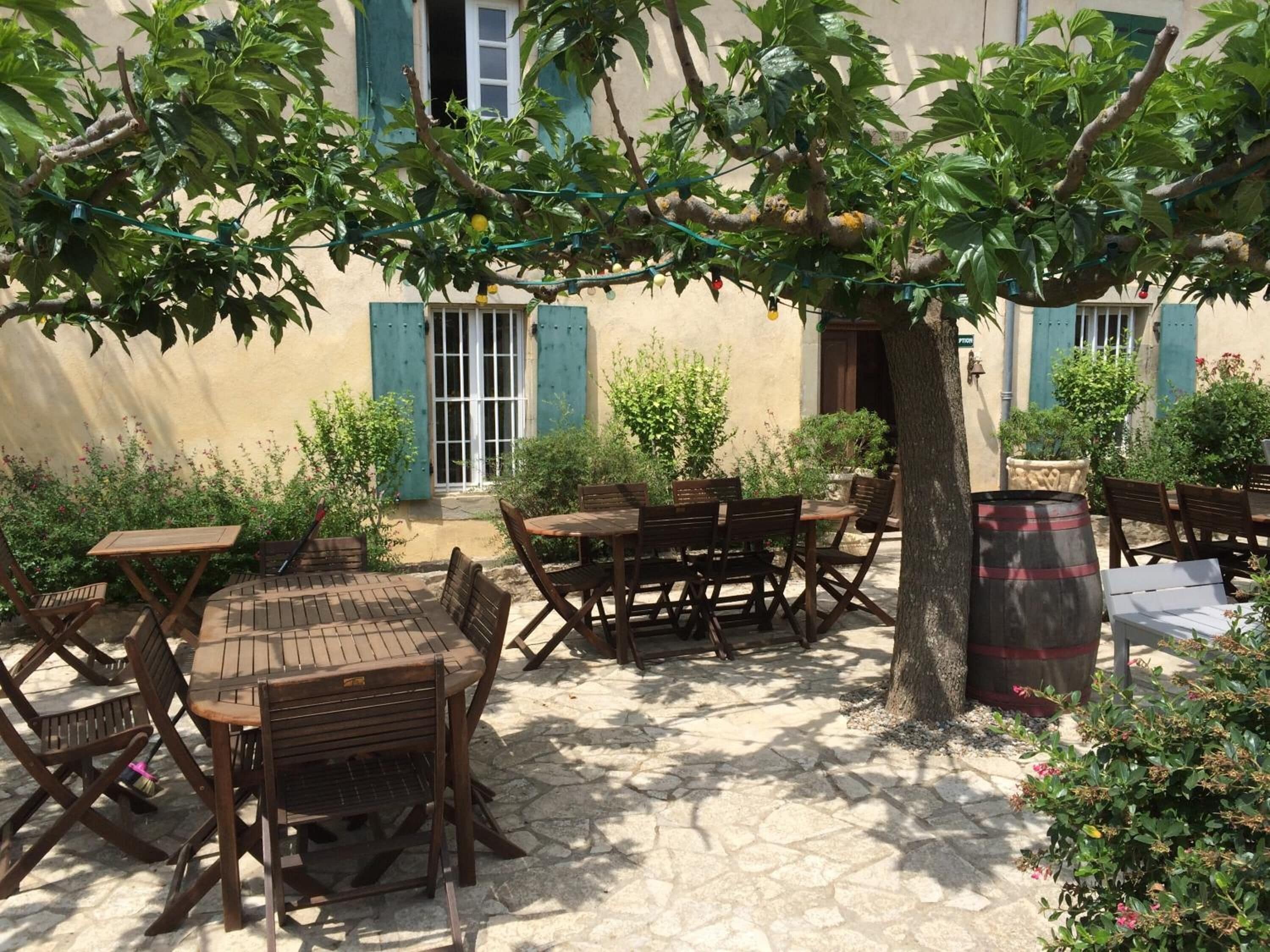 Une terrasse extérieure avec des tables et des chaises en bois, entourée de verdure et bordée d’un bâtiment aux volets bleus.