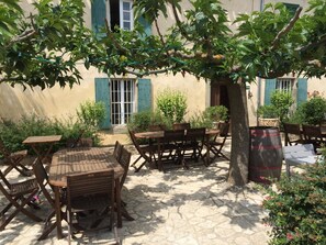 An outdoor patio with wooden tables and chairs, surrounded by greenery and a building with blue shutters. - Domaine de Fraisse (Leuc)