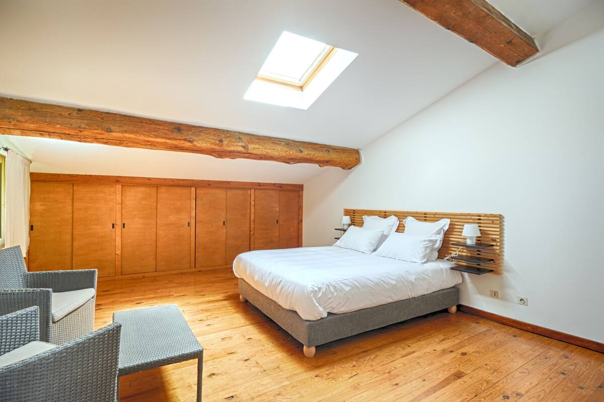 A bedroom with a skylight, wooden wardrobe, bed, bedside table, and wicker chairs.