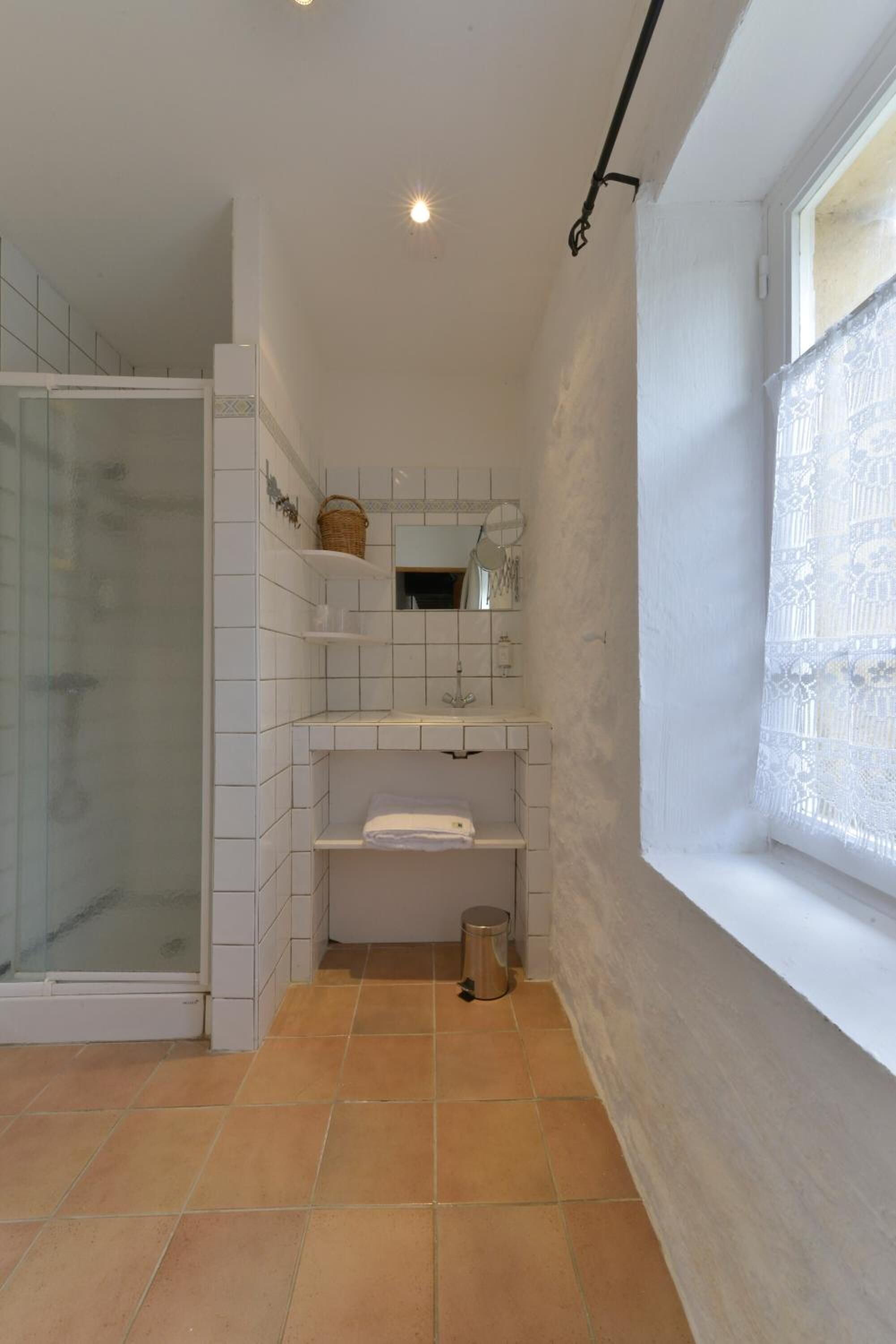 Un bagno con pareti piastrellate di bianco, un box doccia e una finestra con tende in pizzo.