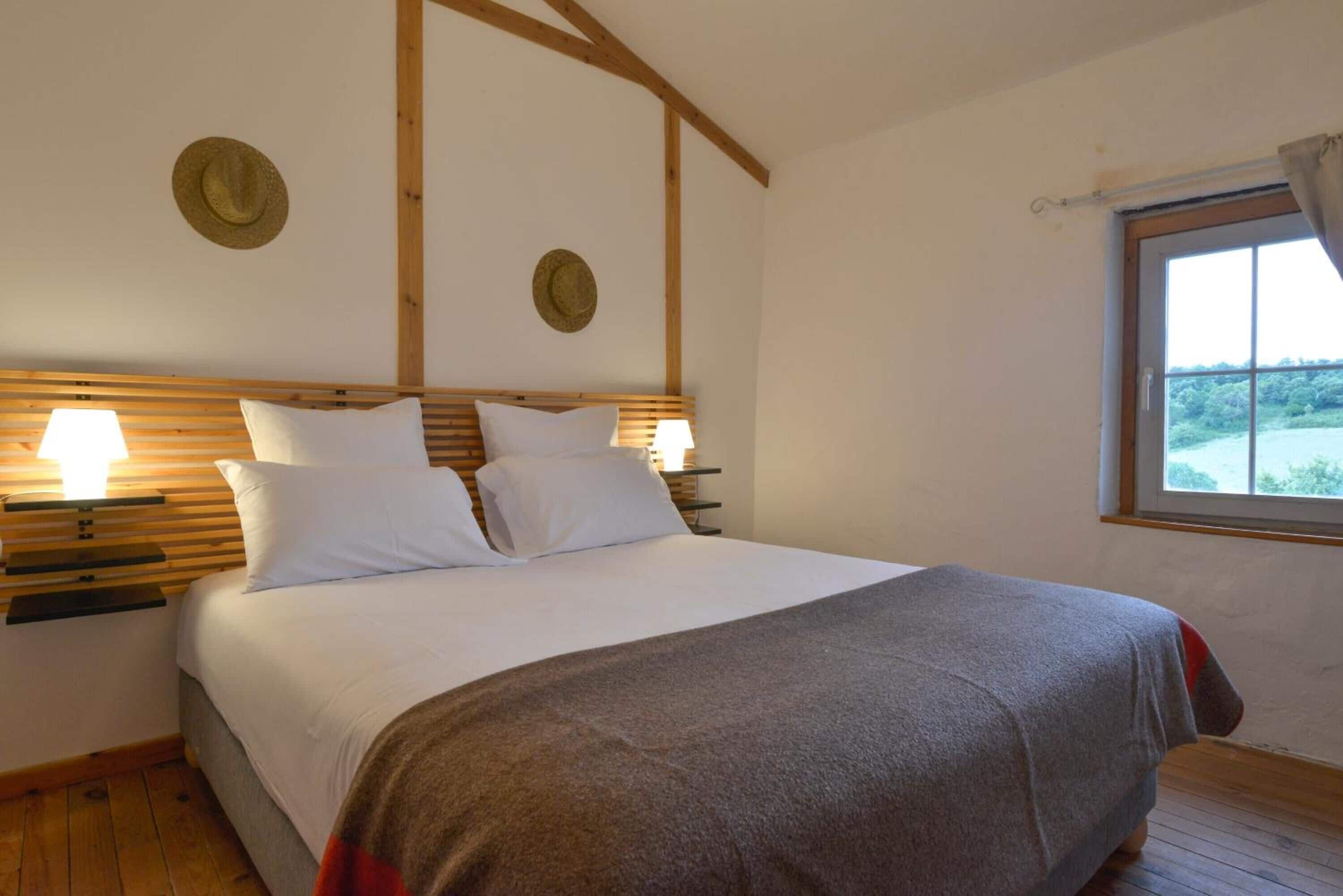 Un dormitorio con cabecera de madera, dos mesitas de noche con lámparas, una cama grande con ropa blanca y una ventana con vistas a zonas verdes.