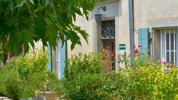 A stone-paved courtyard with a table and chairs, surrounded by lush greenery and a building with blue shutters.