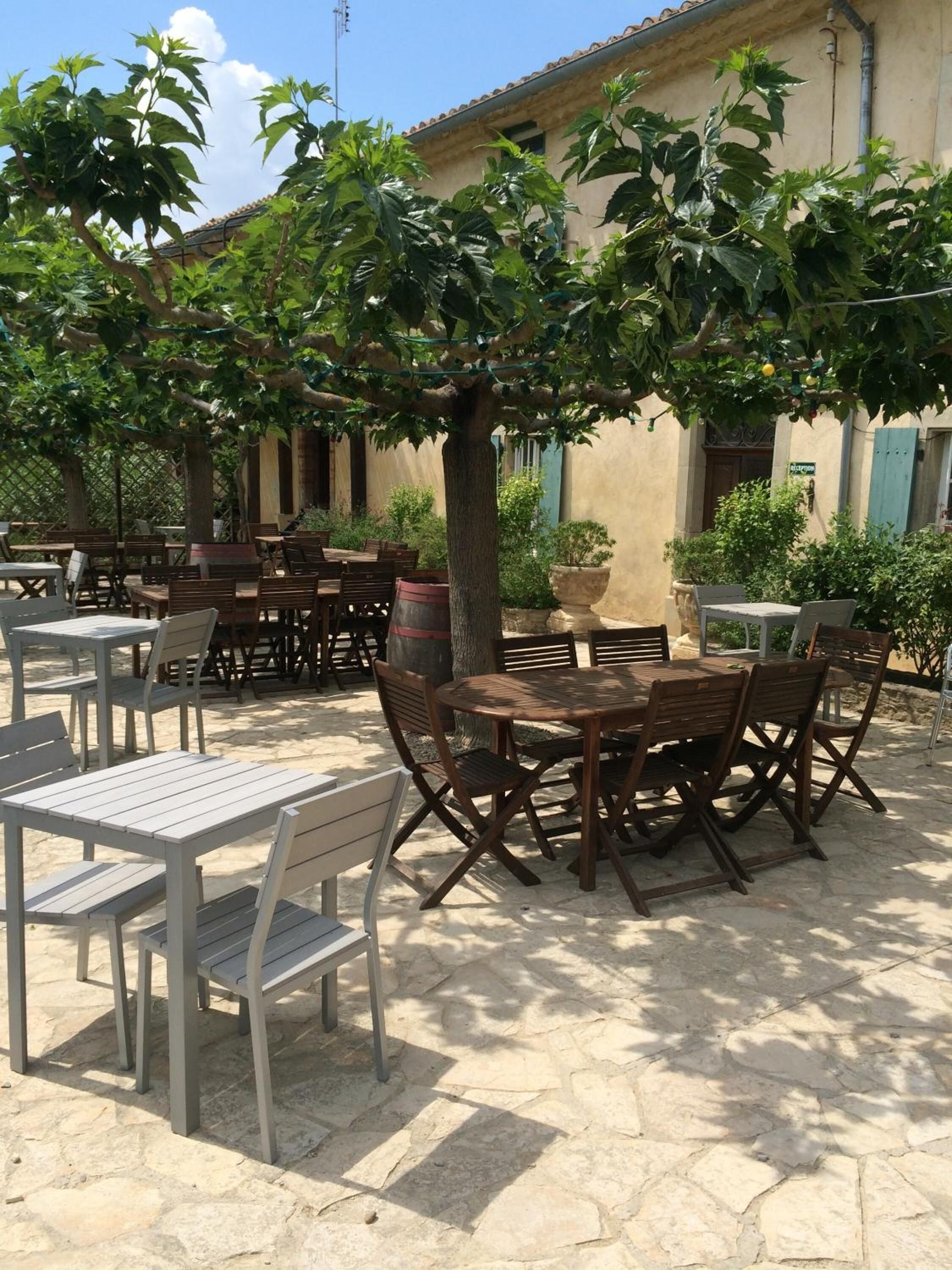 Ein Garten mit Terrassenbereich, Tischen, Stühlen und viel Grün, mit einem Gebäude mit Ziegeldach im Hintergrund.