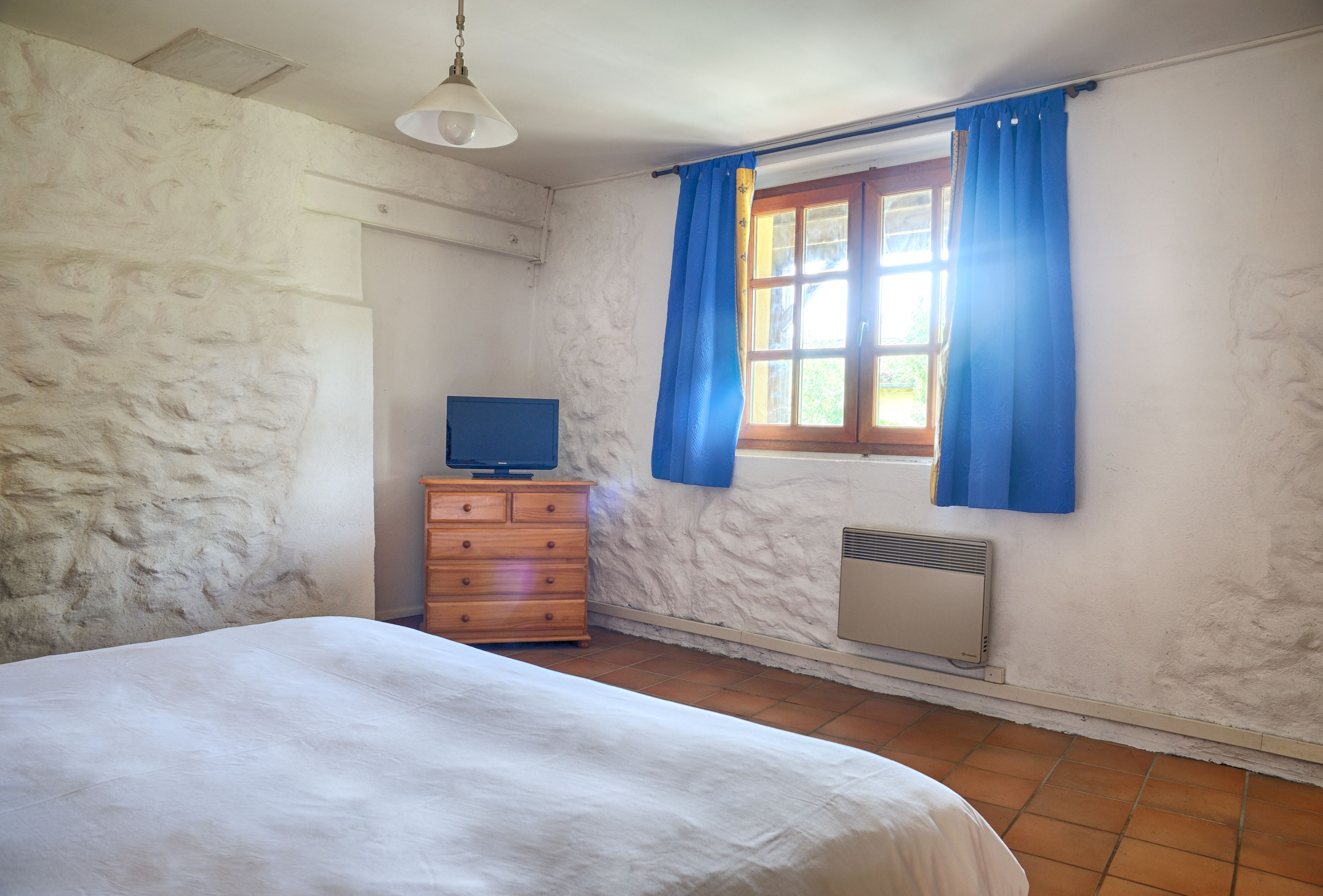 Una camera da letto con un letto, una cassettiera in legno, una televisione, una finestra con tende azzurre e un riscaldatore a parete.