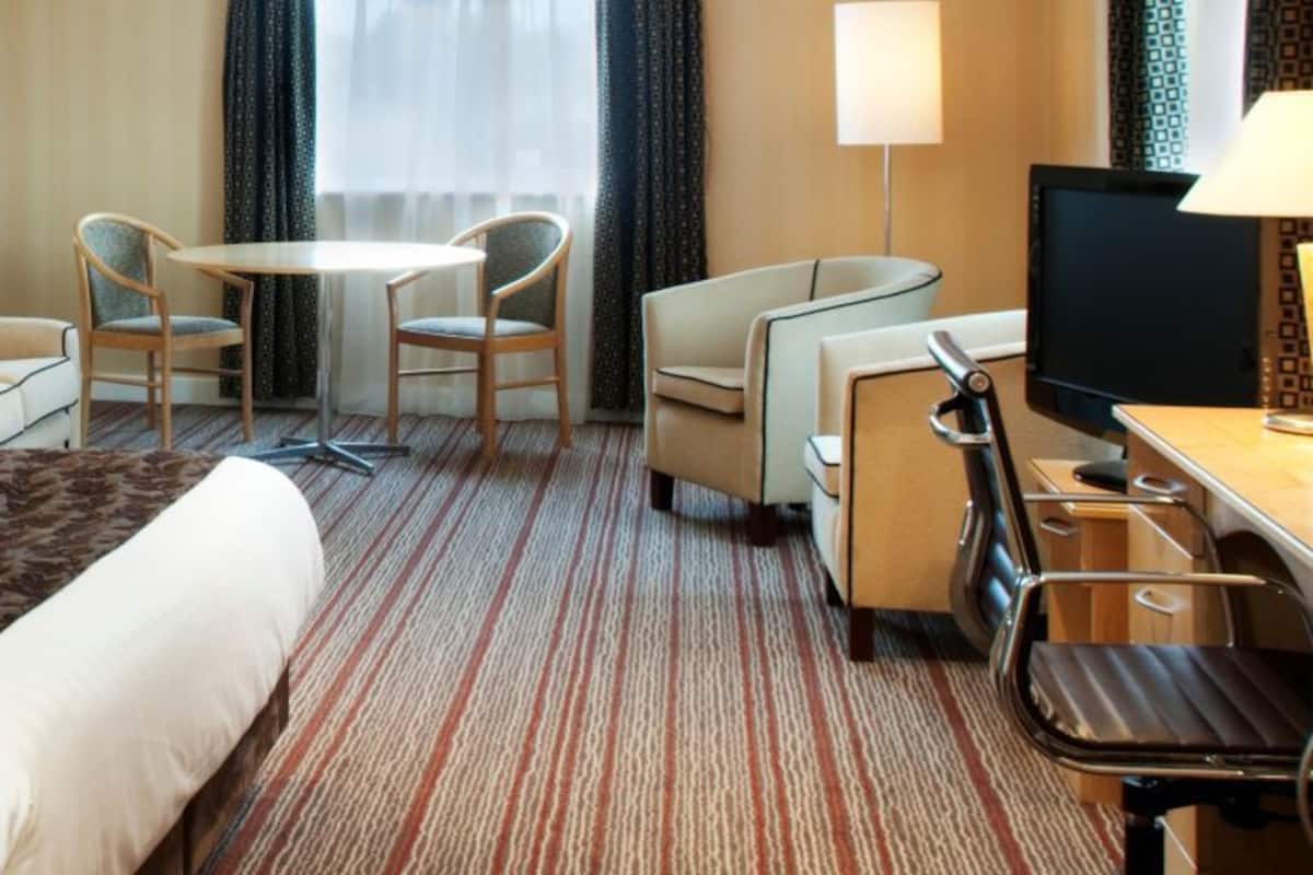 A hotel room with a large bed, a desk with a computer, a small table with chairs, and a window with curtains.