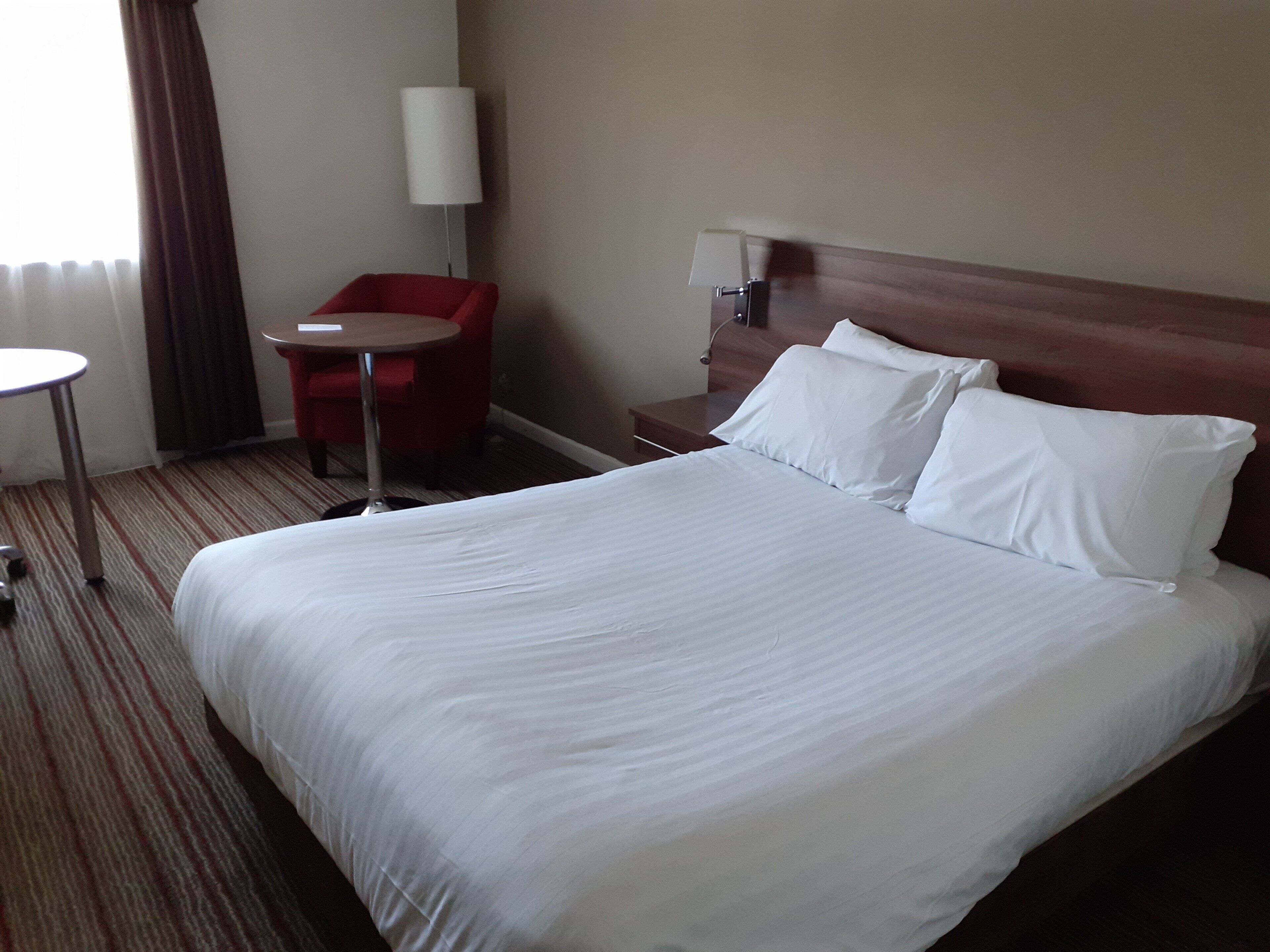 Ein Hotelzimmer mit einem großen Bett, einem kleinen runden Tisch, einem roten Sessel und einer Lampe.