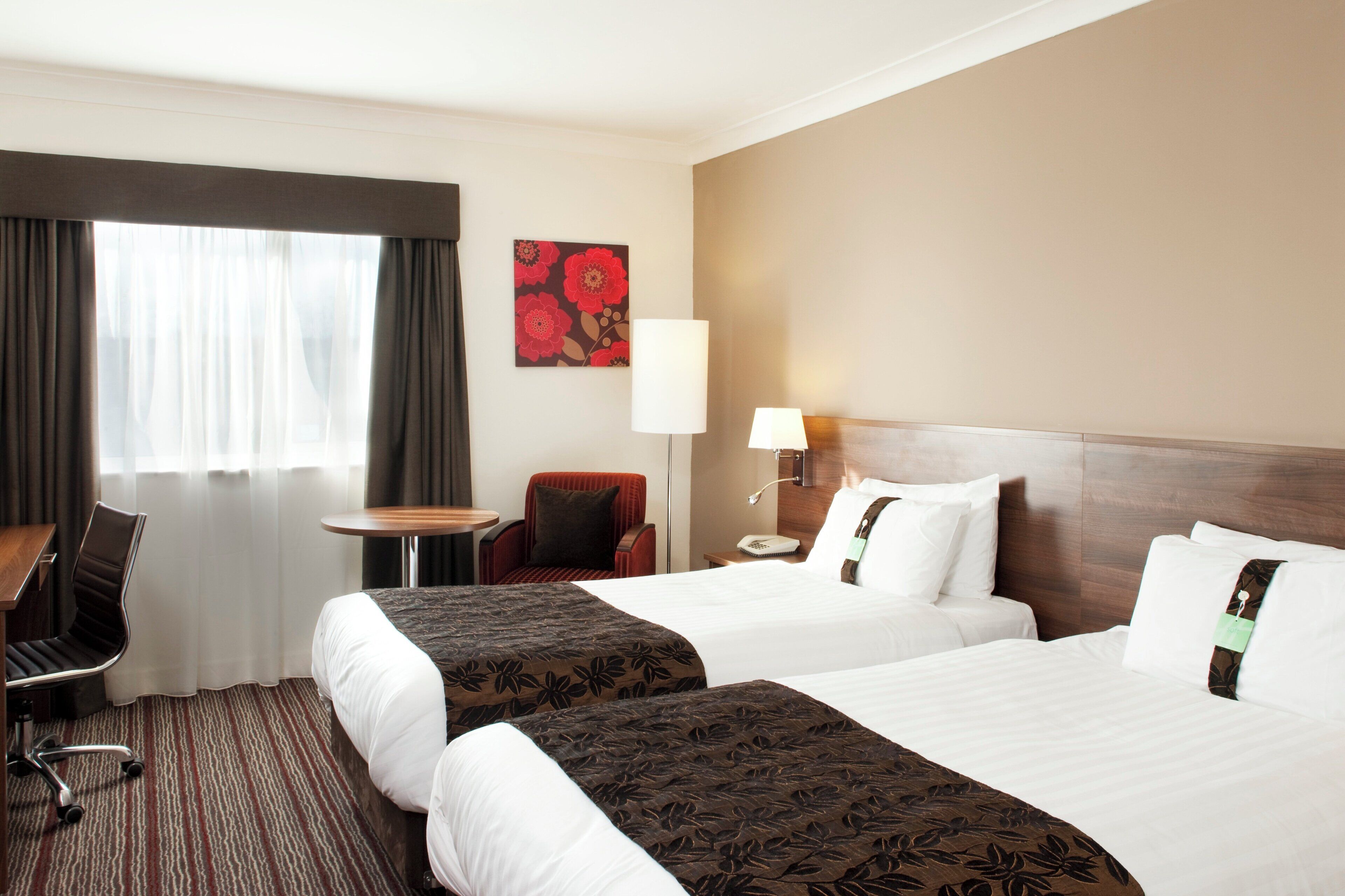 Une chambre d’hôtel avec deux lits, un bureau, une chaise, une lampe et une fenêtre avec des rideaux.