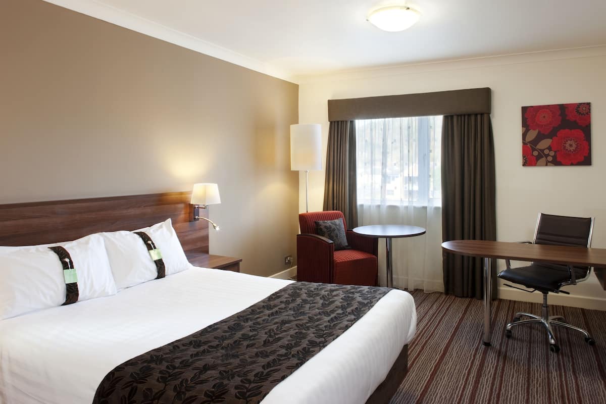 A hotel room with a large bed, a desk, a chair, a small table, and a window with curtains.