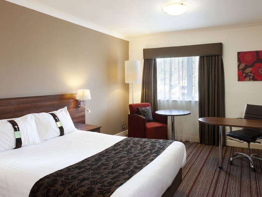 A hotel room with a large bed, a desk, a chair, a small table, and a window with curtains.