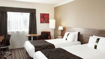 A hotel room with two beds, a desk, a chair, a lamp, and a window with curtains.
