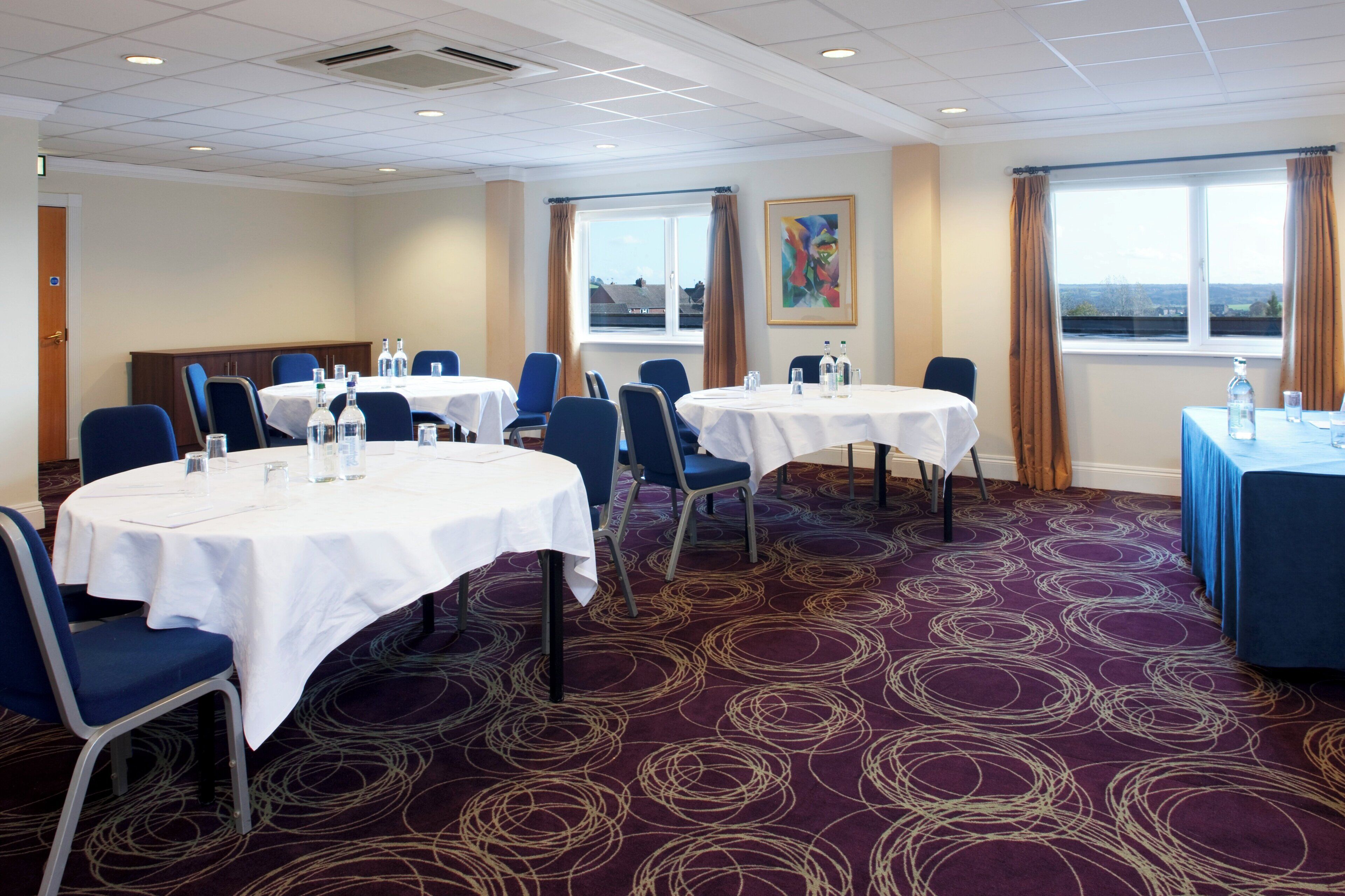 A hotel conference room with round tables, chairs, and a carpeted floor.