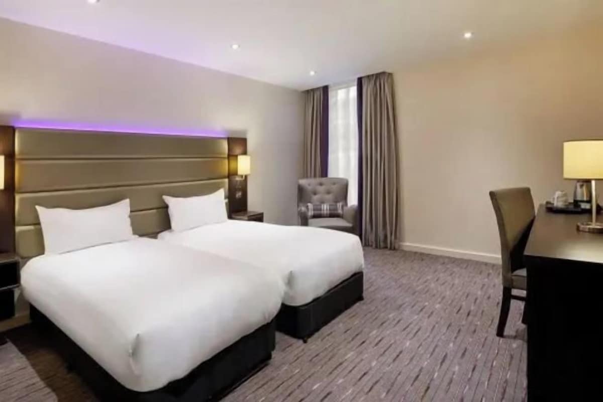 A hotel room with two beds, a desk, a chair, and a lamp.