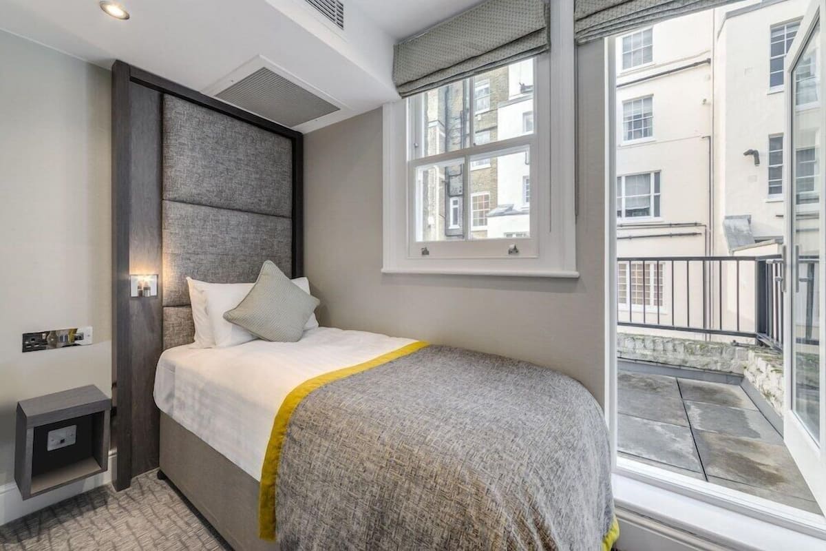 A bedroom with a bed, a bedside table, a window, and a view of buildings outside.
