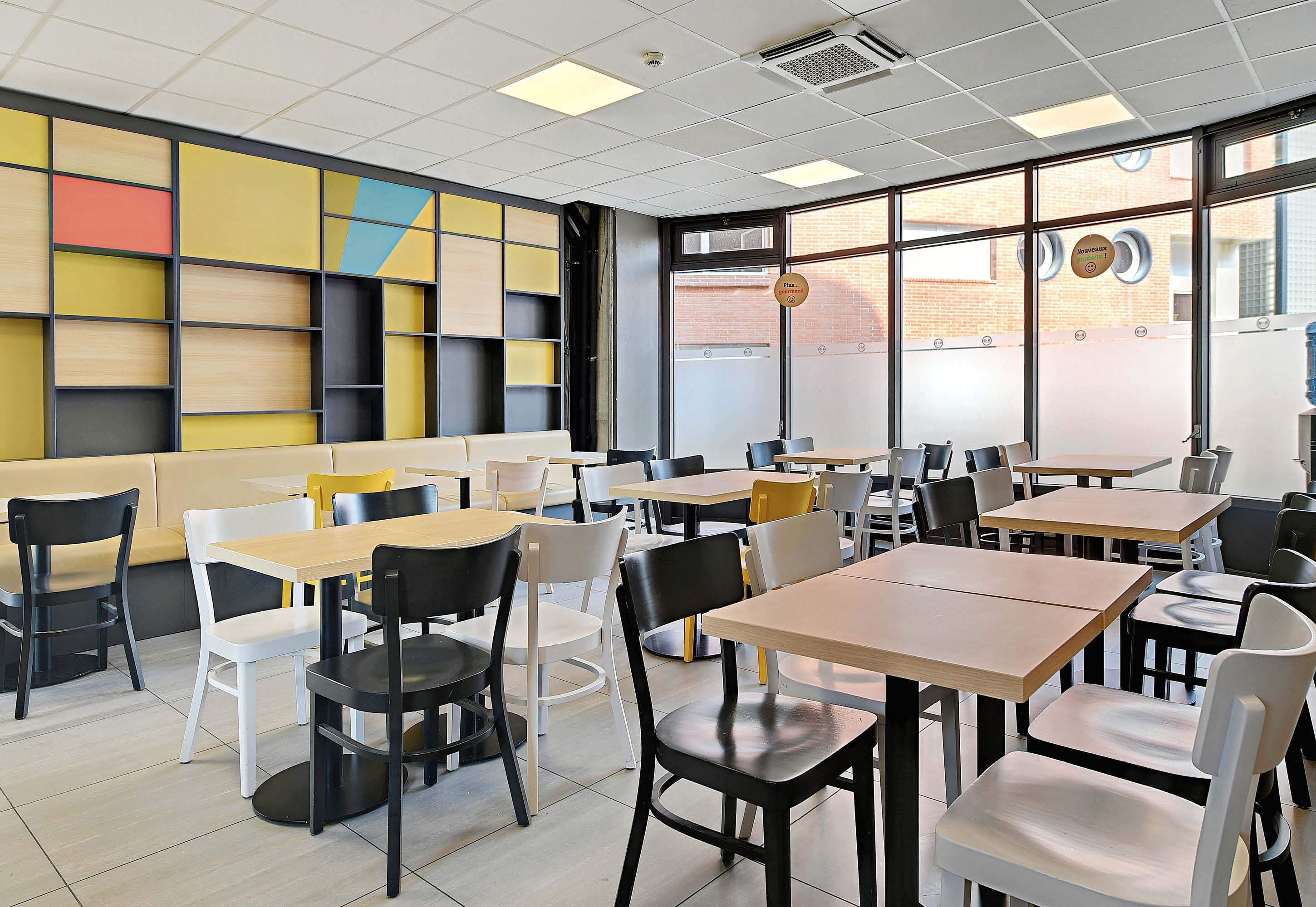 Un café moderno con diversas opciones de asiento, como mesas y cabinas, un diseño de pared colorido y ventanas amplias que aportan luz natural.