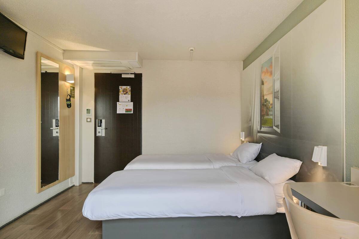 Een hotelkamer met twee bedden, een klein tafeltje en een stoel. Er is een deur die naar een andere kamer leidt, en een wandlamp.
