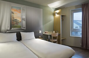 A hotel room with a bed, a desk, a chair, a window with curtains, and a painting of a castle.