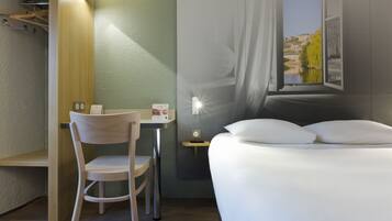 Une chambre d’hôtel comprenant un lit, un bureau avec une chaise, un porte-manteau et une fenêtre donnant sur un immeuble et une étendue d’eau.
