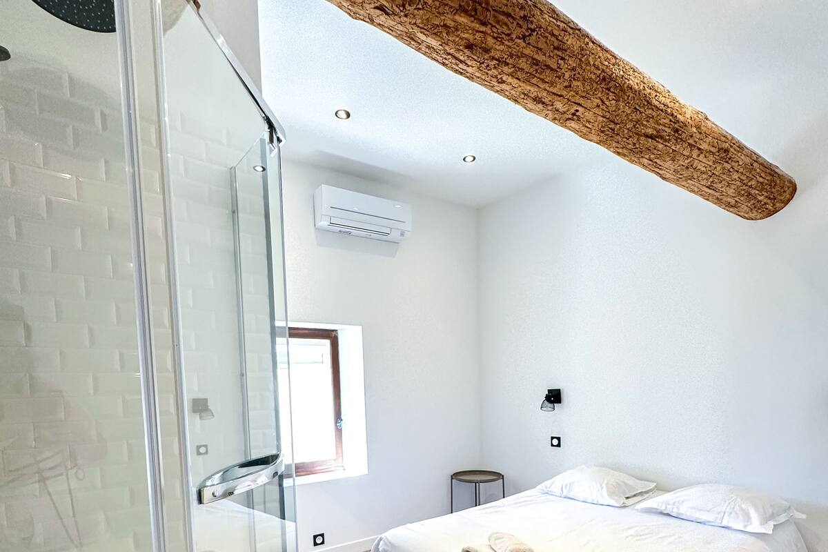 A modern bedroom with a glass shower enclosure, a large bed with white linens, and a wooden ceiling beam.