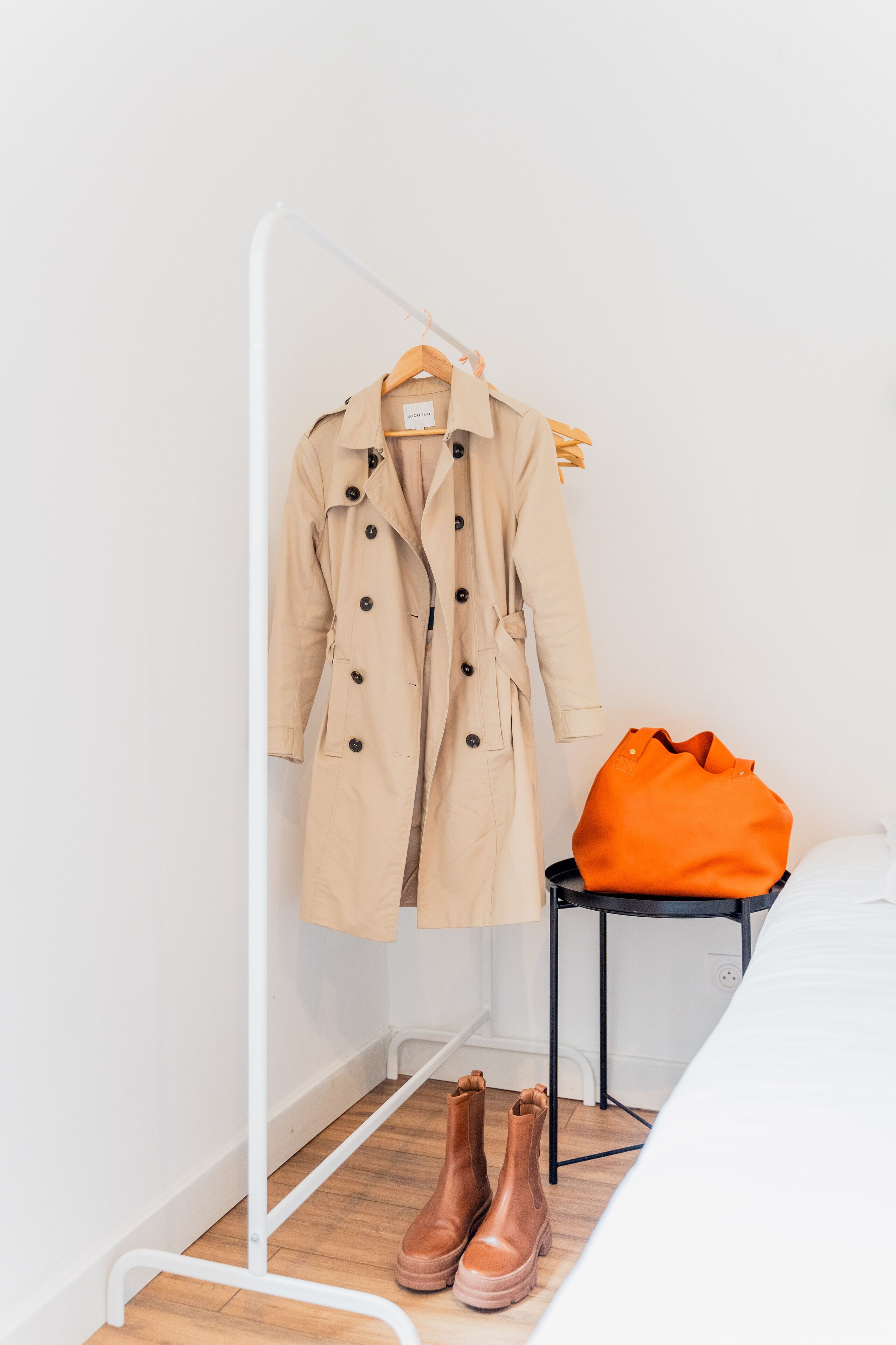Et værelse med en seng, et knagered med en beige trenchcoat, en orange taske og et par brune støvler.