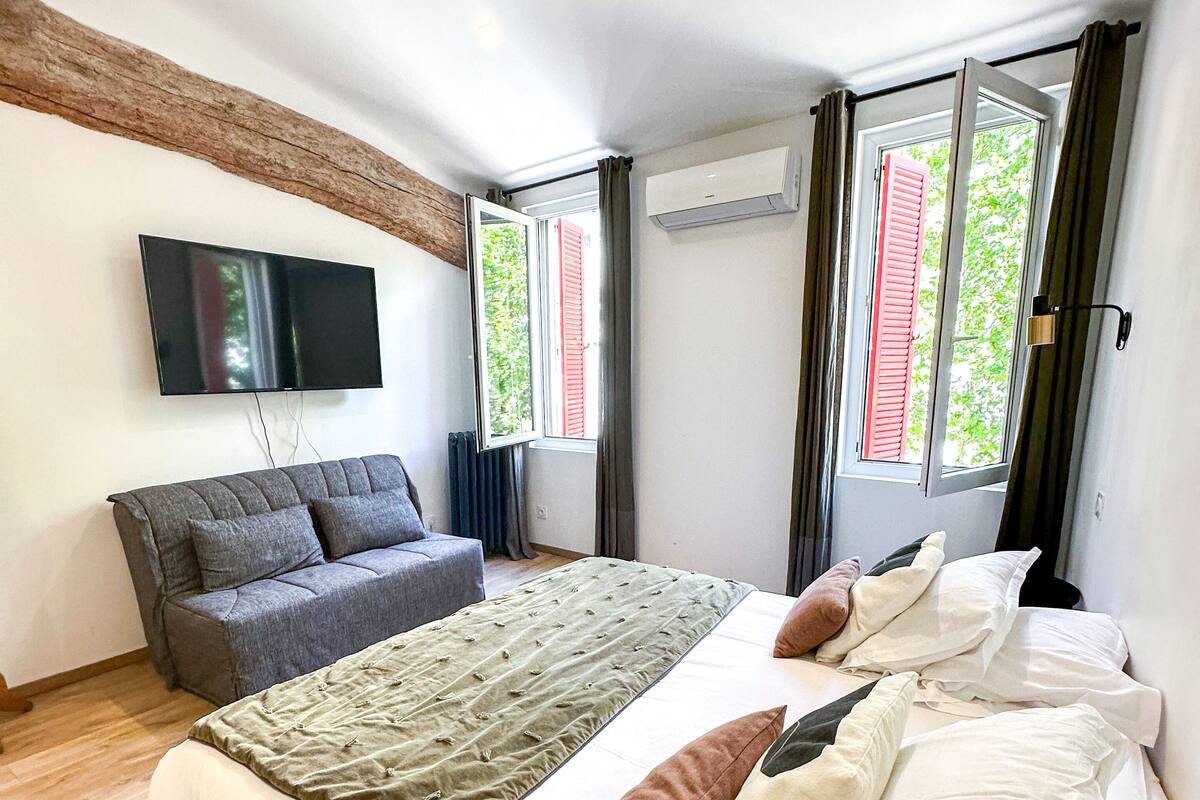 A modern bedroom with a large bed, a grey sofa, a flat-screen TV mounted on the wall, and a window with shutters.