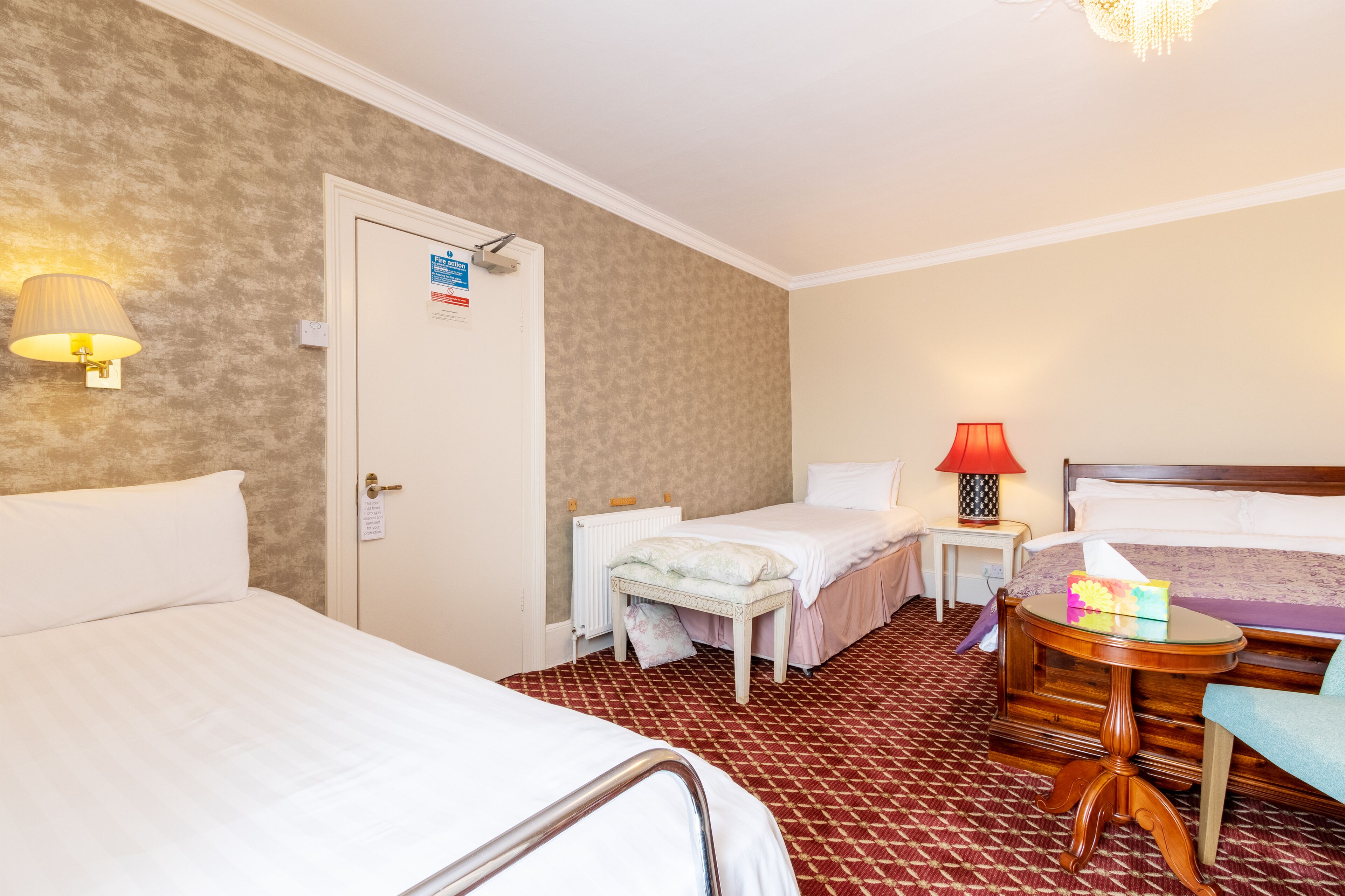 Hotellihuone, jossa on kaksi sänkyä, pieni pöytä lampun kanssa ja punakuvioinen matto.