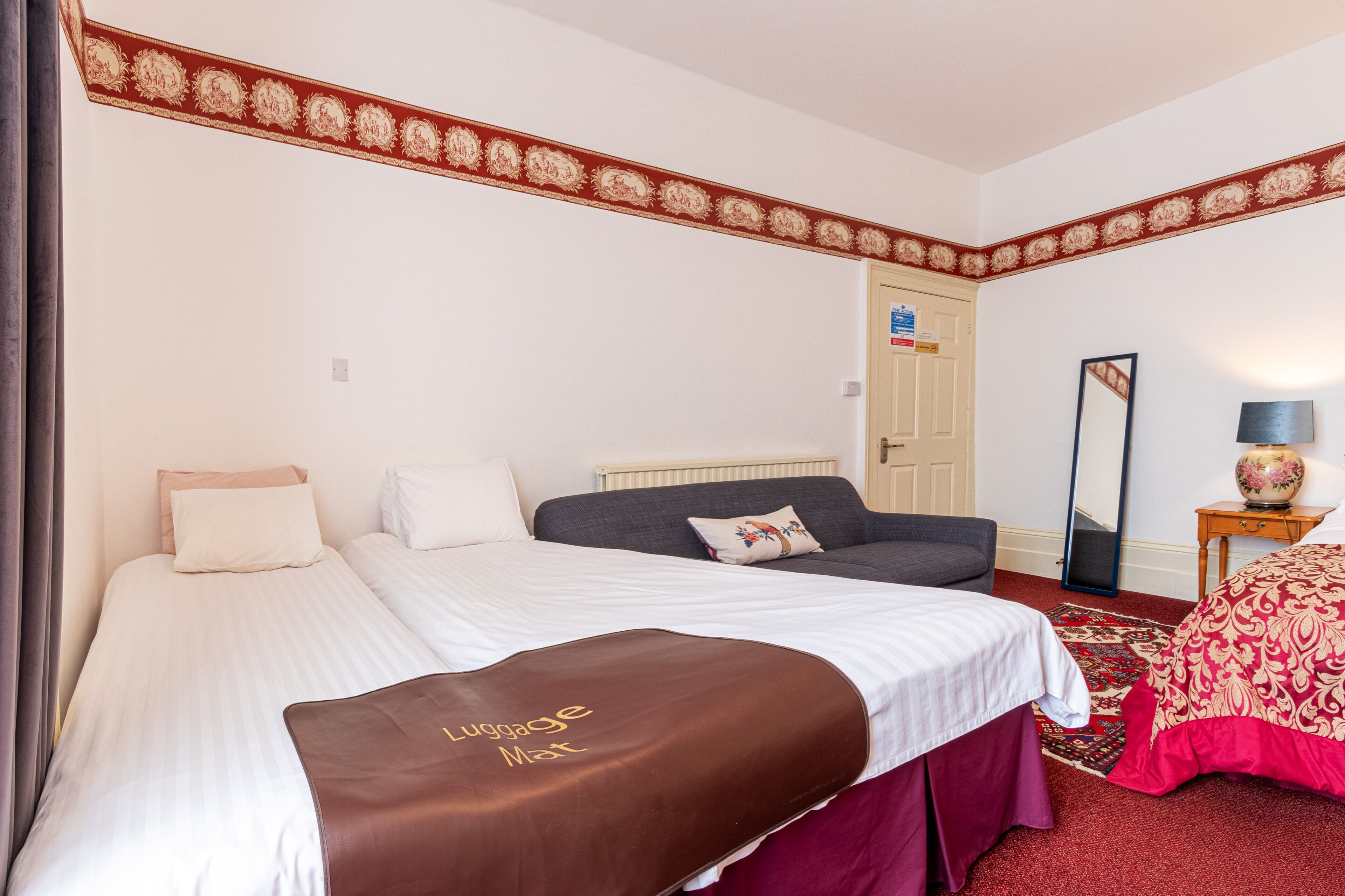 Habitación de hotel con dos camas, un borde rojo y blanco en el techo, un espejo y una mesita con una lámpara.
