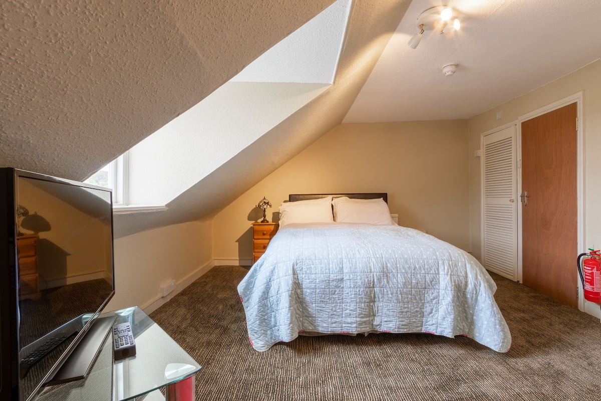 A bedroom with a bed, a desk with a laptop, a TV, and a window.