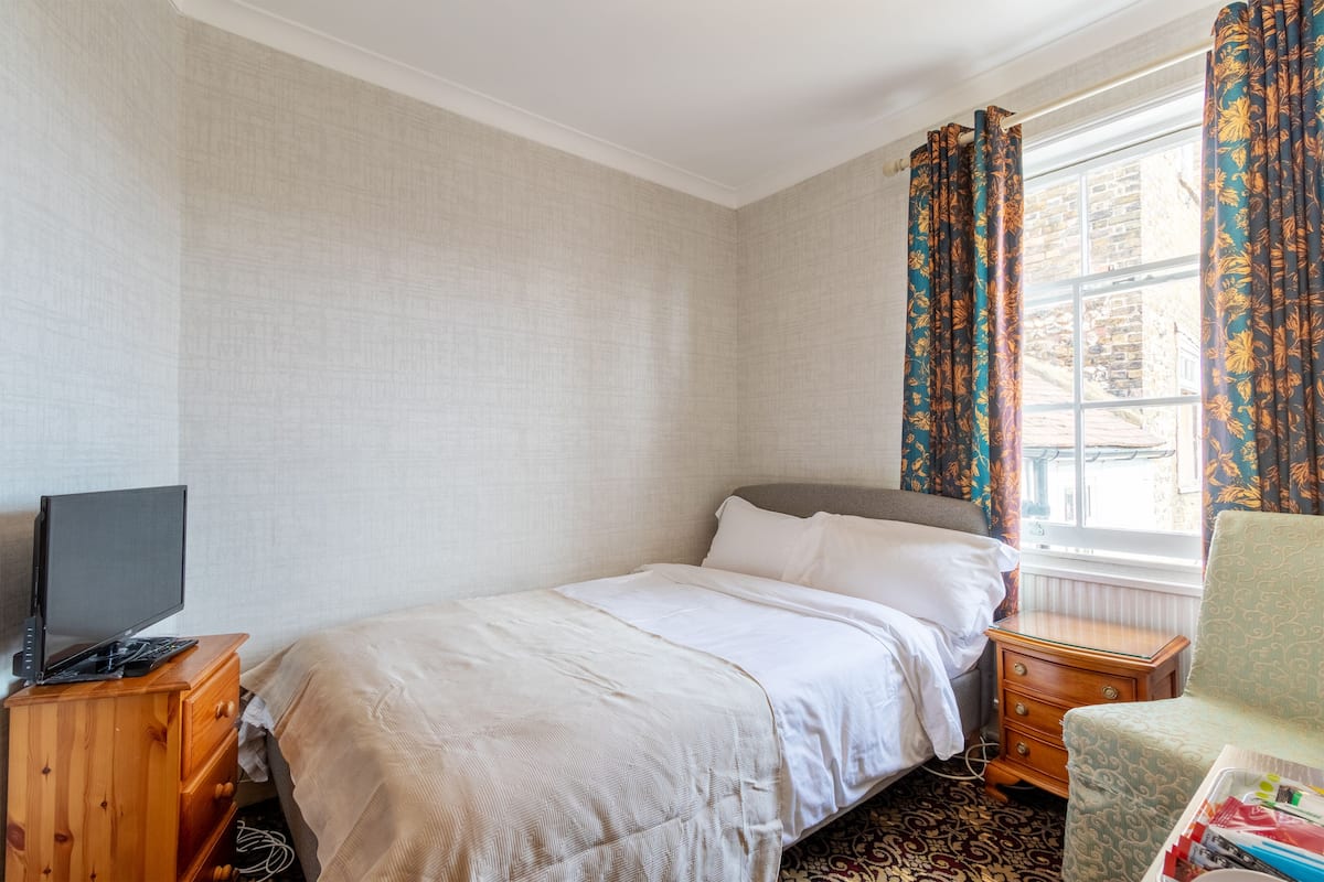 A bedroom with a bed, a wooden nightstand, a television on a dresser, and a window with curtains.