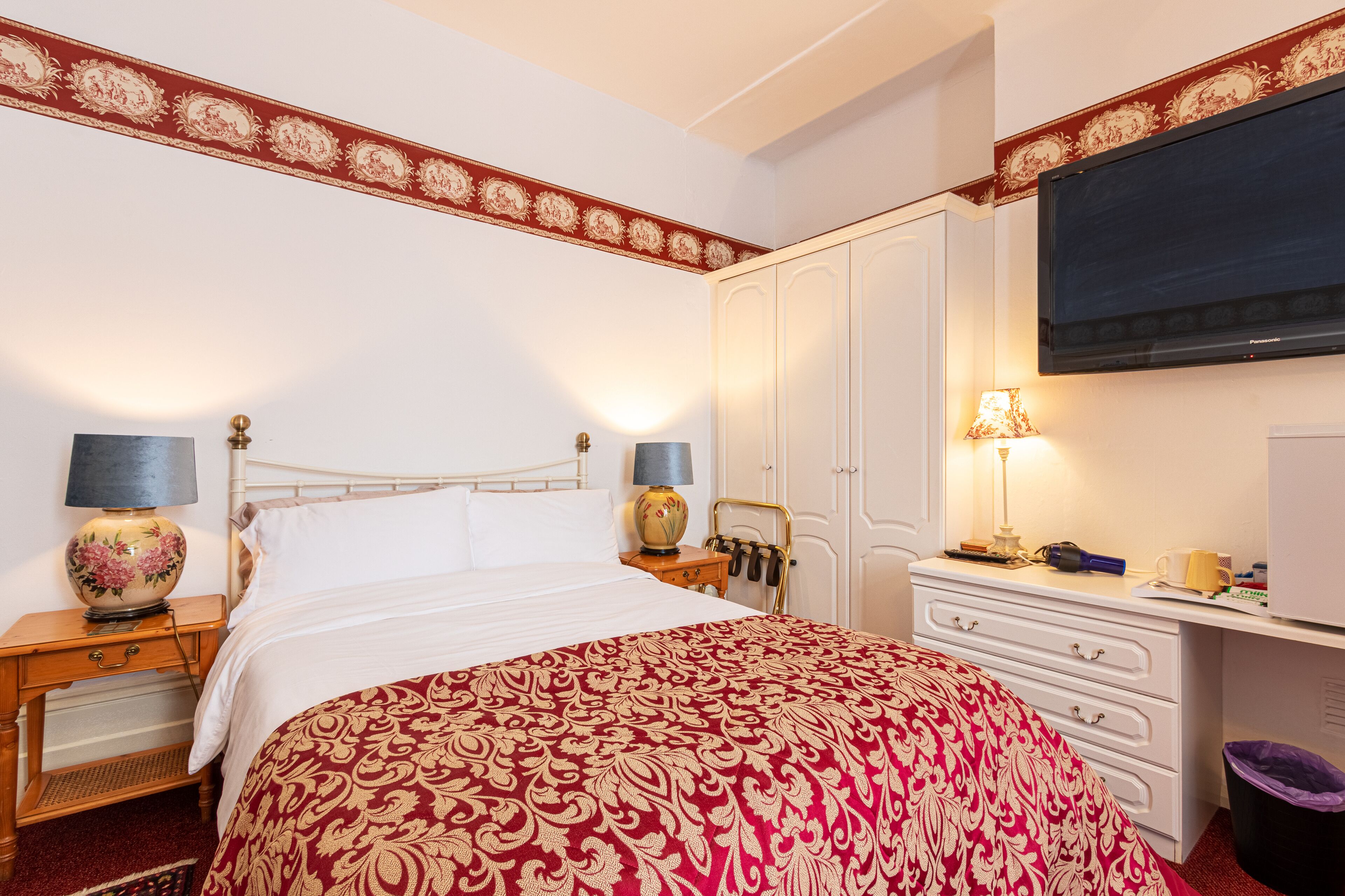 A hotel room with a large bed, two bedside tables with lamps, a wardrobe, a desk with a television, and a trash bin.